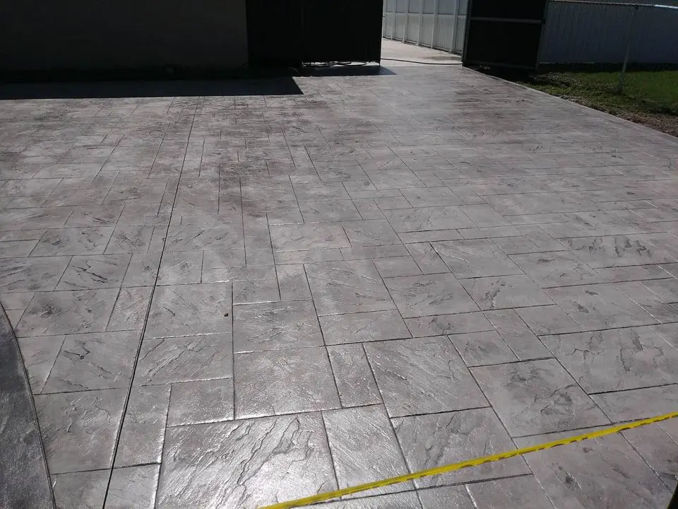 A large concrete patio, crafted by a leading concrete contractor in Albuquerque, features a stamped pattern resembling tiles. The glossy surface is marked by yellow caution tape in the foreground, while structures and a fence outline the background, showcasing both commercial and residential finesse.