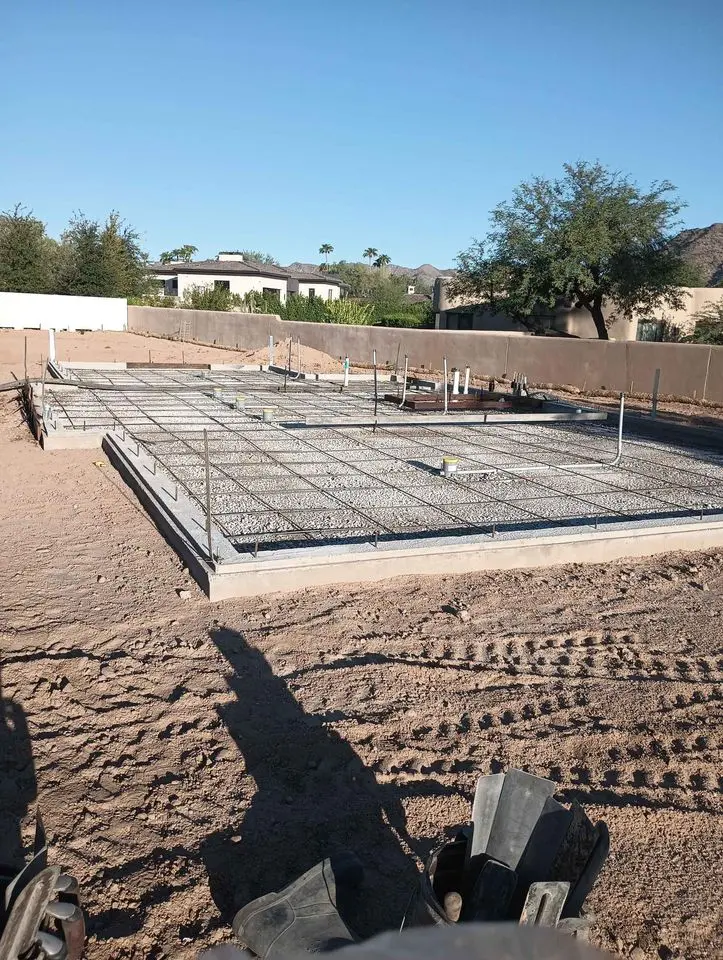 A construction site with a residential concrete foundation grid laid out on a sandy lot. The structure is surrounded by a low wall, with trees and a house visible in the background under a clear blue sky. Various construction tools are scattered in the foreground.