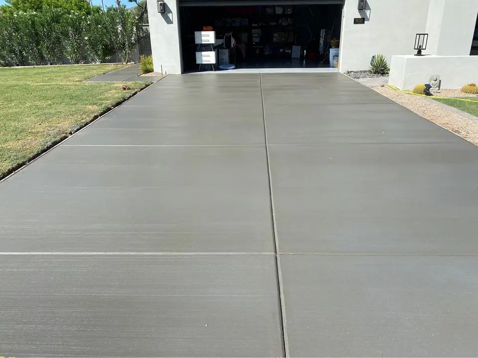 A smooth concrete driveway crafted by Quality Concrete NM leads to an open garage with lawn on the left and a gravel border on the right. Various items and tools fill the shelves inside, basking in Albuquerque NM's sunlight that casts shadows across the driveway.