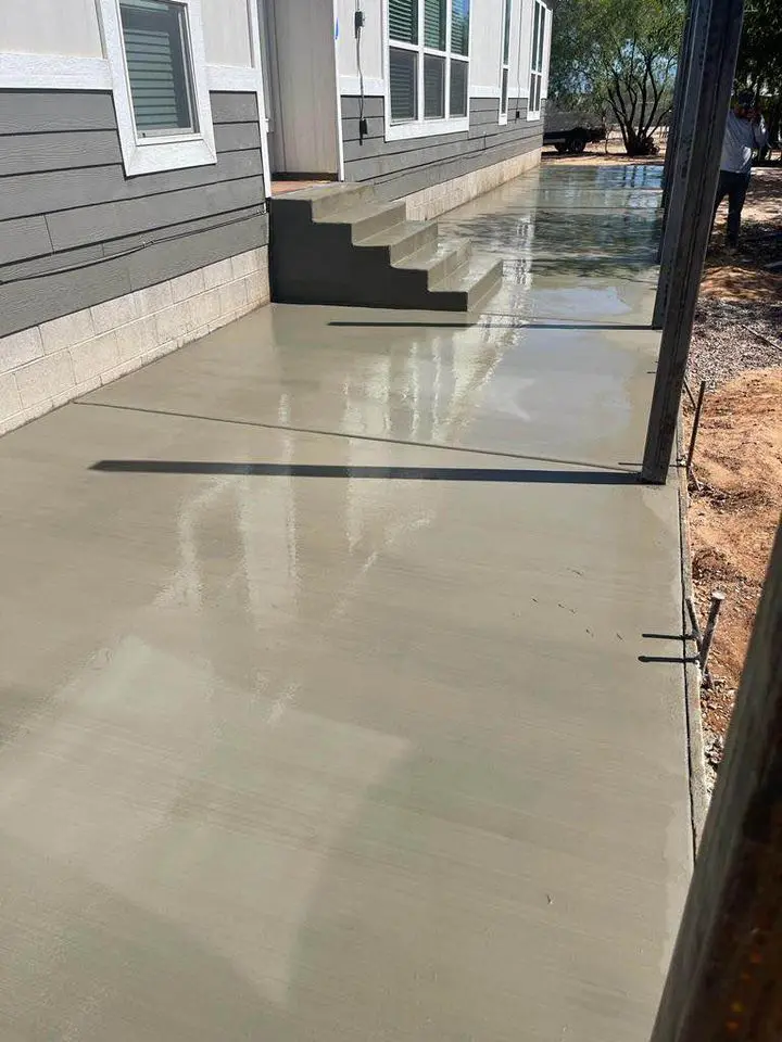 Freshly poured by Quality Concrete NM, the smooth and wet concrete pathway and stairs lead elegantly to a building's entrance in Albuquerque, NM. A person is partially visible in the background near trees, subtly complementing the scene crafted by expert concrete driveway contractors.