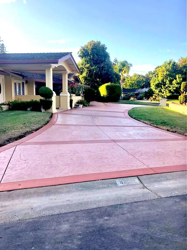 A house with a red-tinted concrete driveway, expertly crafted by Quality Concrete NM, is bordered by green grass and well-trimmed bushes, leading to a carport. The driveway curves slightly under the Albuquerque NM sky, framed by trees and a clear blue background.