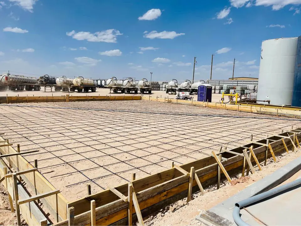 A construction site in Albuquerque, NM, showcases a large rectangular area prepared for pouring by a commercial concrete contractor. The space is outlined with wooden planks and reinforced with a metal rebar grid. Trucks stand ready under a blue sky with scattered clouds, ensuring quality work by Quality Concrete NM.