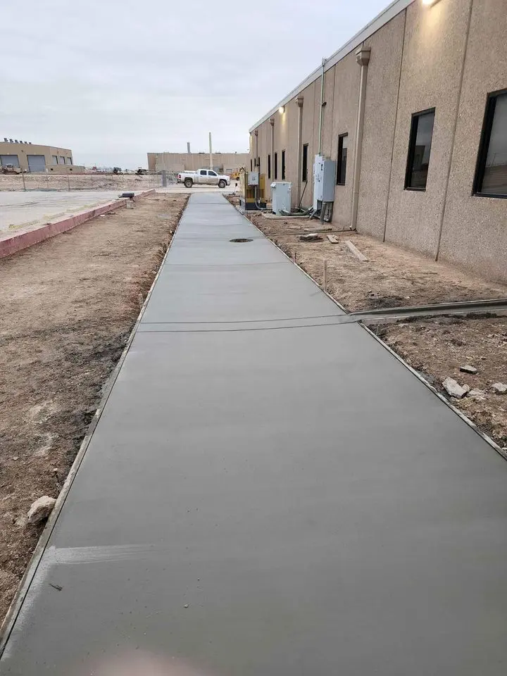 A newly paved concrete sidewalk, crafted by a quality concrete contractor in Albuquerque, NM, runs alongside a beige building in a construction area. The sky is overcast, and loose soil lines the path. A parked truck and unfinished structures add to the scene's industrious atmosphere.