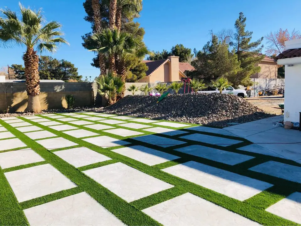 A landscaped yard in Albuquerque features a grid pattern of concrete slabs and artificial grass. Tall palm trees and a pile of rocks decorate the backdrop alongside a white building to the right. Designed by a local concrete contractor, this patio shines under the sunny, clear blue sky.