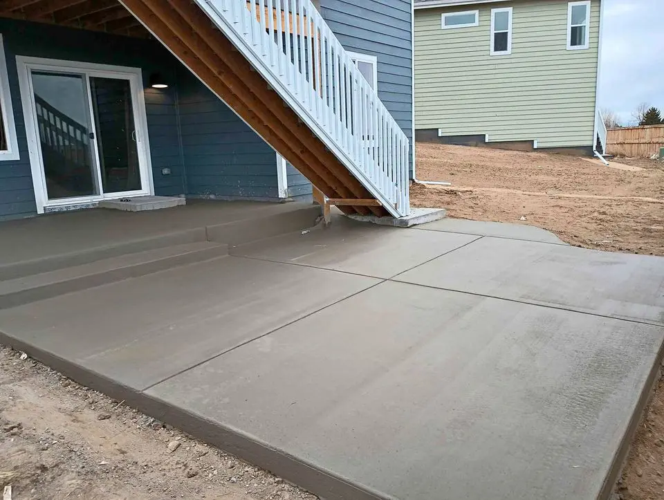 Newly poured concrete patio crafted by a skilled concrete contractor, with steps leading to a raised deck. Attached to a house with blue siding and white trim, it showcases another house with green siding in the background on a grassy lot, capturing the essence of Albuquerque, NM.