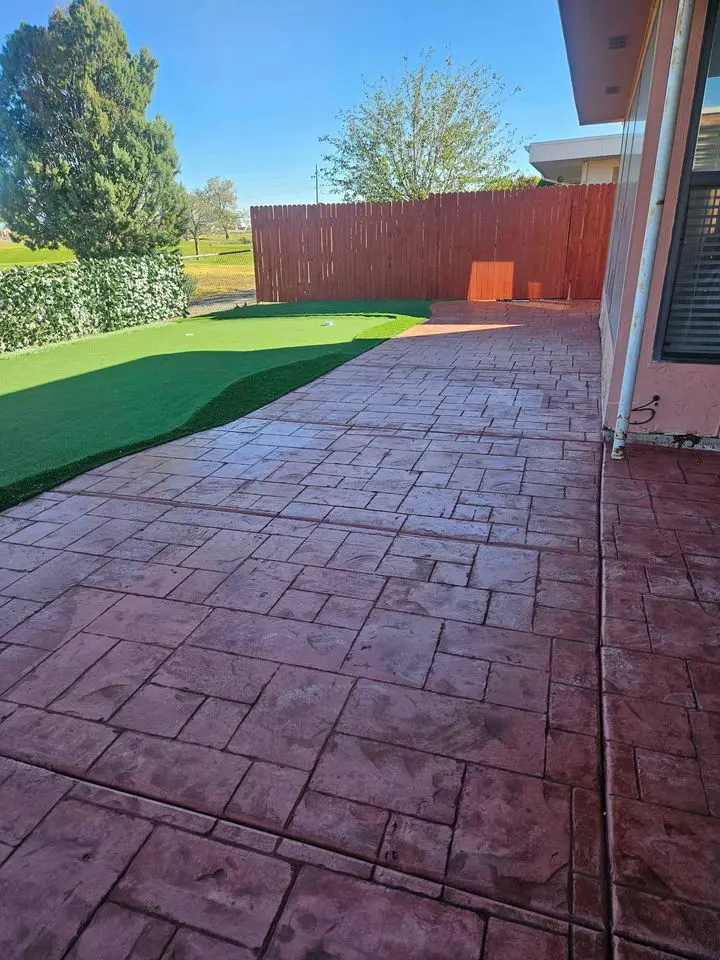 A backyard in Albuquerque, NM, showcases a red brick-pattern patio beside artificial grass. A tall wooden fence and lush greenery grace the yard, all under a clear blue sky. This inviting space was expertly crafted with care by a skilled concrete contractor.