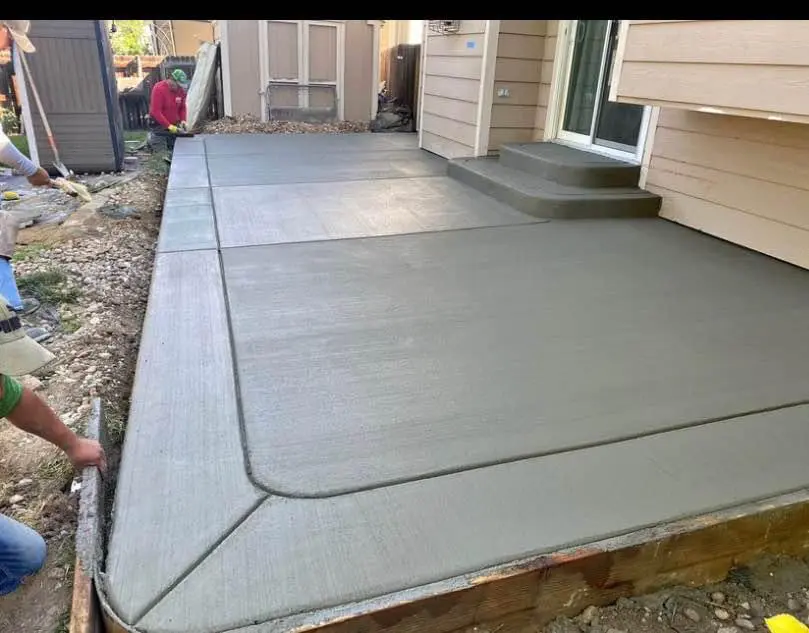 A group of people in Albuquerque, NM, are smoothing freshly poured concrete on a backyard patio next to a house. The concrete contractor ensures distinct sections and expertly shapes the edges. The patio leads up to steps at the house entrance, creating an elegant transition.