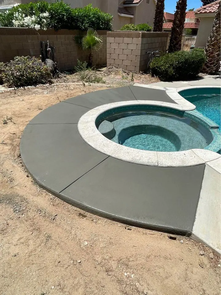 A circular outdoor hot tub seamlessly connects to a swimming pool, nestled in an Albuquerque oasis with a sleek concrete pool deck. The newly laid gray surface by expert concrete contractors complements the dry landscaping, while lush plants and trees frame the scene against a block wall backdrop.