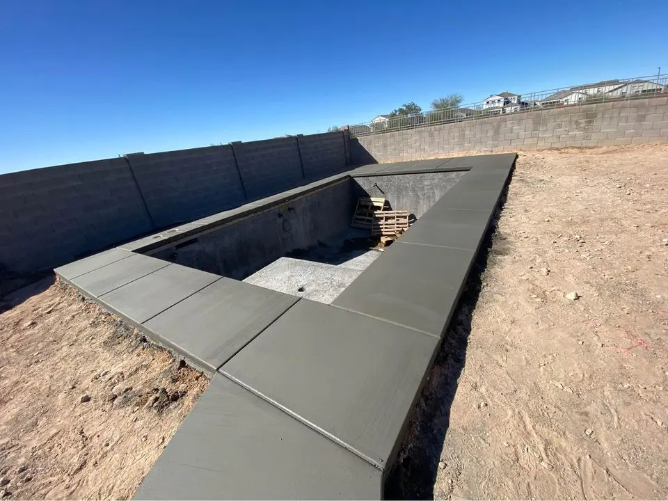 A rectangular in-ground pool under construction boasts a sturdy concrete shell and edge, crafted by skilled concrete contractors, and is nestled in a sandy Albuquerque yard. The empty pool has wooden pallets inside, while a tall brick wall encloses the space under a clear blue sky.