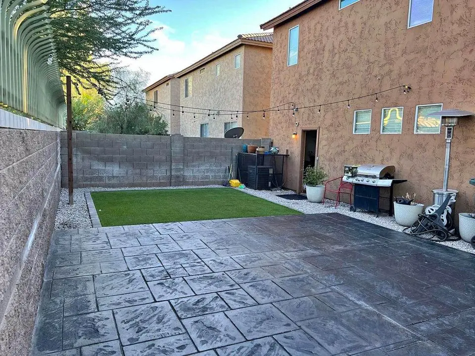 A backyard in Albuquerque with a concrete patio featuring stamped designs, a small lawn area, string lights overhead, a grill, and planters along the wall. The yard is enclosed by a cinder block fence, with two houses visible in the background.