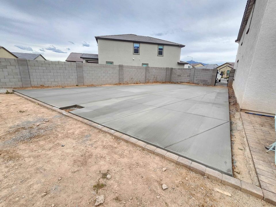 A freshly poured concrete slab, crafted by a skilled concrete contractor in Albuquerque, fills a backyard area surrounded by a low brick wall. The ground is bare, and two houses are visible in the background under an overcast sky. Contact us for a free quote on your next project!