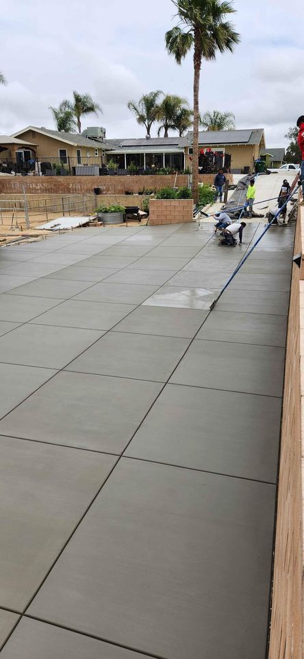 Workers smooth out freshly poured residential concrete on a large driveway. Palm trees and small houses are visible in the background under a cloudy sky, as the Concrete Contractor Albuquerque expertly sections the driveway into large square slabs.