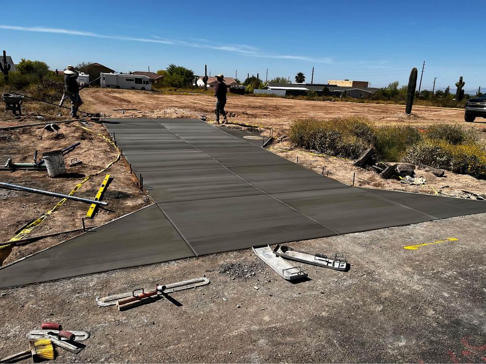 Two workers from Quality Concrete NM are paving a new residential concrete path in a desert-like area, surrounded by sand and shrubs. The clear blue sky stretches above, with construction tools scattered around the site.