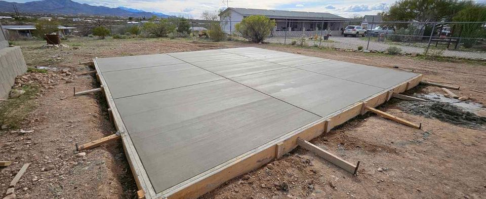 A freshly poured concrete slab in Albuquerque, NM, divided into even sections, rests in a dirt area with mountains and houses visible in the background. Wooden forms border the slab as part of a concrete contractor's craftwork under the partly cloudy sky.