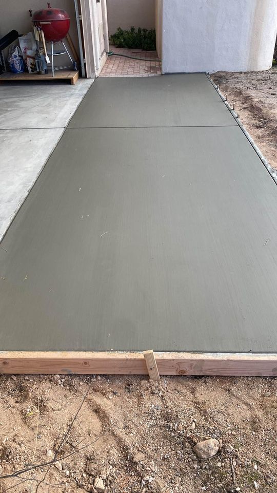 Freshly poured residential concrete slab on the ground, bordered by wooden planks, next to a garage. A red grill and various tools are visible in the garage in the background. The area around the slab is dirt and gravel.