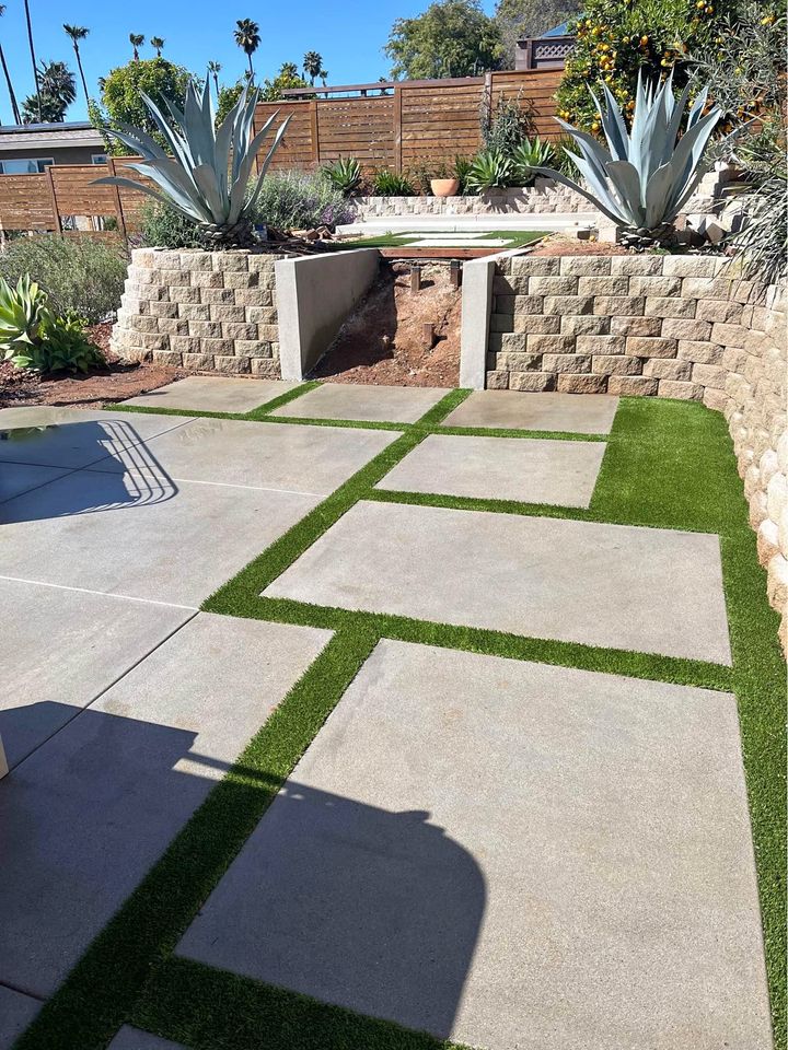 Outdoor patio featuring large stone tiles and strips of lush artificial grass. A raised garden bed filled with succulents and agave plants sits in the background, complemented by a built-in circular fire pit. A privacy fence stands under a clear blue sky, crafted by expert concrete contractors in Albuquerque.