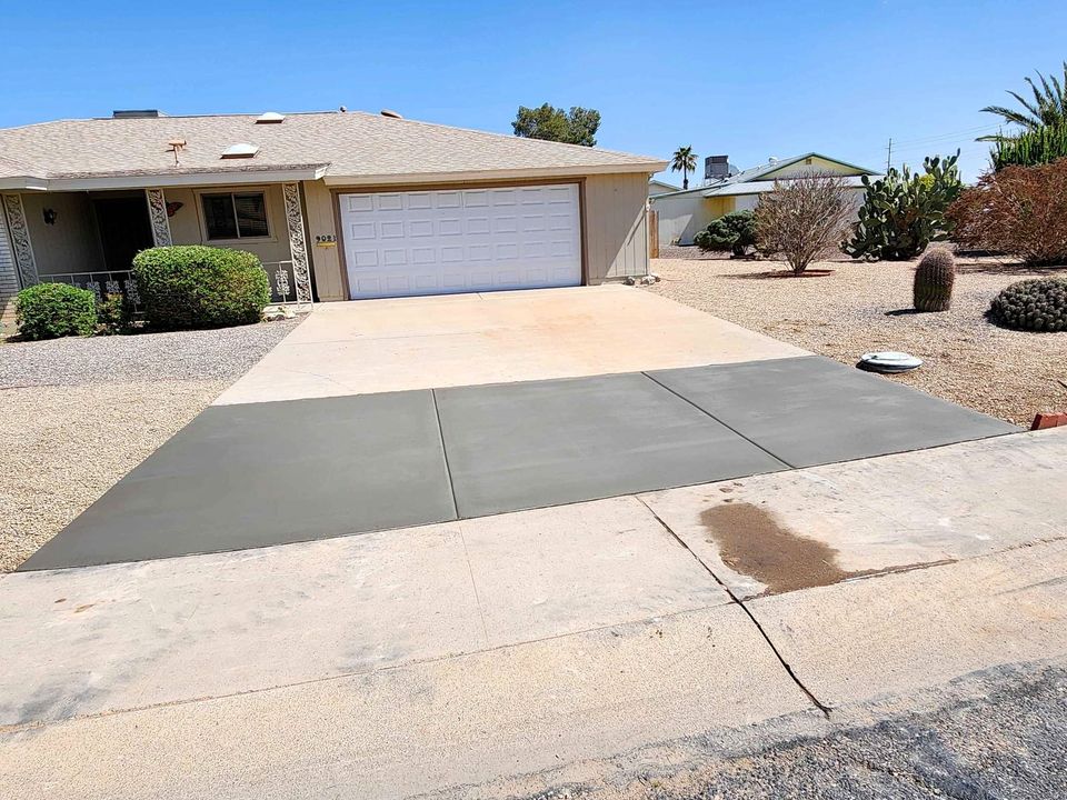 A residential house with a two-car garage and a newly paved driveway showcases expertly laid foundations. The driveway smoothly blends into the sidewalk, freshly finished by a top Concrete Contractor in Albuquerque. The landscaped front yard features rocks, bushes, and small trees under a clear blue sky.