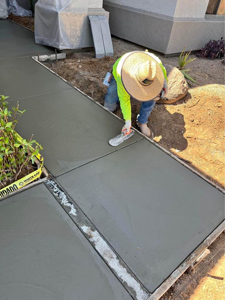 A construction worker in a green shirt and straw hat is expertly smoothing freshly poured concrete on a sidewalk with a trowel. Surrounded by dirt, plants, and caution tape, this concrete contractor from Albuquerque ensures quality work in both commercial and residential projects.