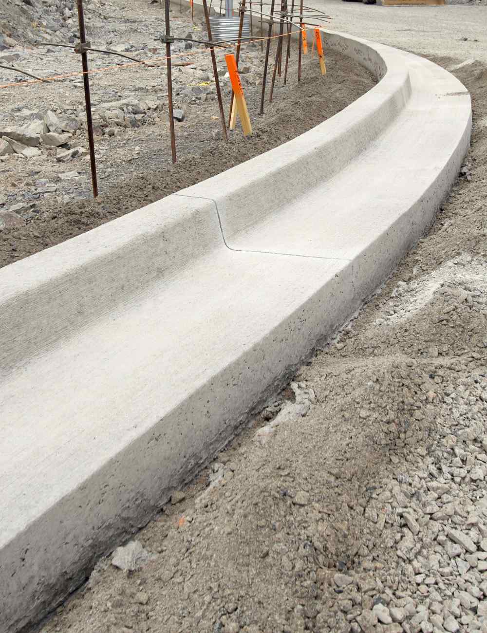 A newly constructed concrete curb runs along a dirt pathway, with wooden stakes and bright orange markers in the background. This Albuquerque construction site showcases the skill of the contractor, with visible gravel and soil adding to the scene's dynamic landscape.