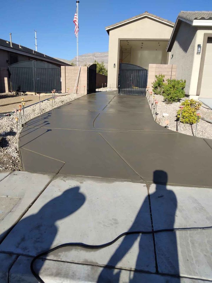 A freshly laid concrete driveway, expertly crafted by Quality Concrete NM, leads to a gated garage of a house in Albuquerque. The surrounding area features gravel and small shrubs. Two shadows of people appear on the pavement in the foreground, with an American flag visible in the background.