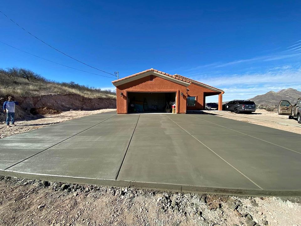 A wide, freshly paved concrete driveway by Quality Concrete NM leads up to a single-story, red brick house with an open garage. Two people stand on the left, and a black SUV is parked on the right under Albuquerque's clear blue sky.