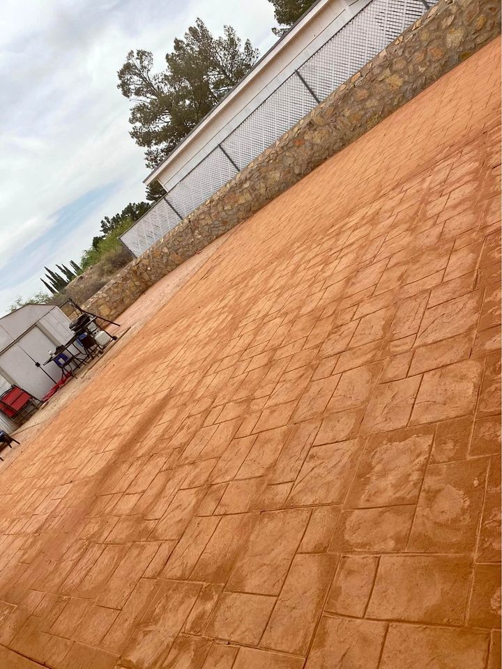 A large, textured, reddish-brown paved area crafted by Quality Concrete NM in Albuquerque features a stone wall and metal fence. Trees and a cloudy sky enhance the view, with outdoor furniture and equipment nestled near the wall.