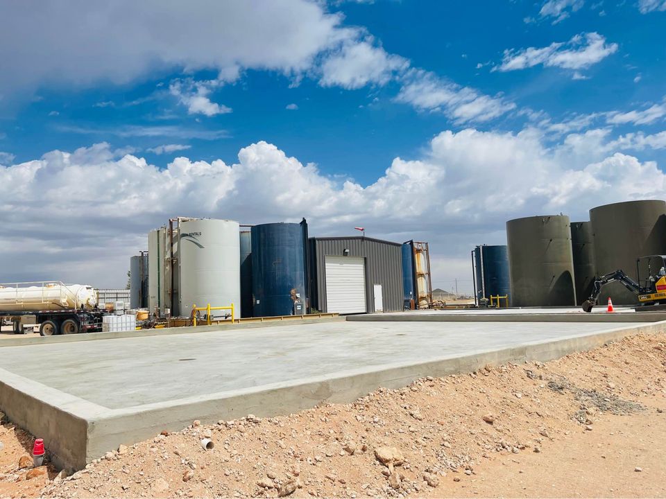 An industrial site sits under a blue sky with clouds, featuring large storage tanks and a small building. In the foreground, a flat concrete area extends, hinting at the handiwork of a Quality Concrete NM expert. Nearby are a truck and construction equipment, framed by Albuquerque NM's distinct rocky terrain.