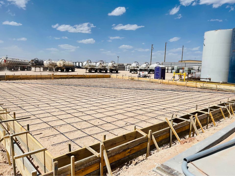 A construction site in Albuquerque, NM, showcases a large rectangular area prepared for pouring by a commercial concrete contractor. The space is outlined with wooden planks and reinforced with a metal rebar grid. Trucks stand ready under a blue sky with scattered clouds, ensuring quality work by Quality Concrete NM.