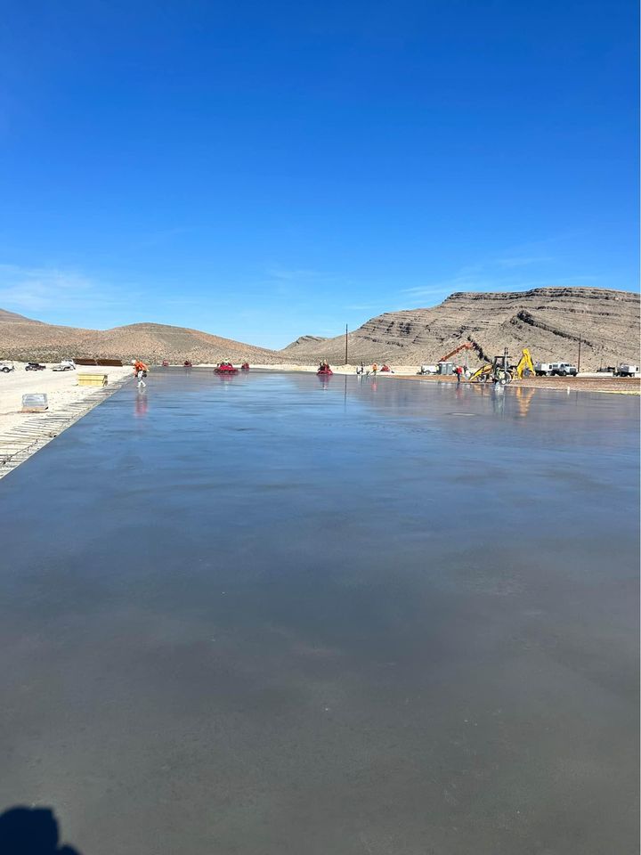 Industrial site with a Quality Concrete NM pad in the foreground, surrounded by large cylindrical storage tanks. A metal building with an open door is in the background, and tanker trucks are parked nearby. The sky over Albuquerque NM is clear and blue.