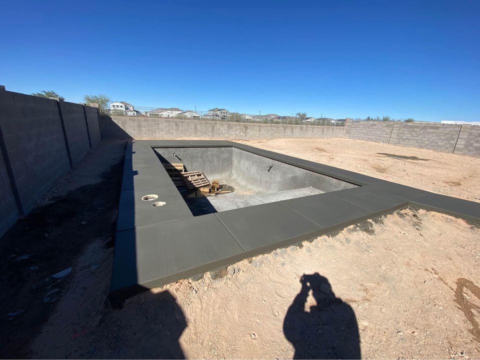 In Albuquerque, NM, the construction site of an unfinished swimming pool awaits completion by skilled concrete contractors. The empty concrete pool is surrounded by dirt in a fenced backyard with a ladder inside. In the foreground, a shadow of the person taking the photo adds to the scene's intrigue.