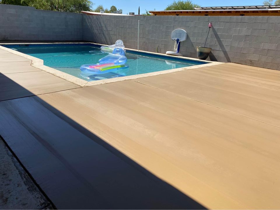 A backyard swimming pool in Albuquerque features a clear inflatable float and a basketball hoop in the corner. The pool is surrounded by a concrete deck, expertly crafted by local concrete contractors, while a high grey wall ensures privacy as the sun casts shadows across the deck.