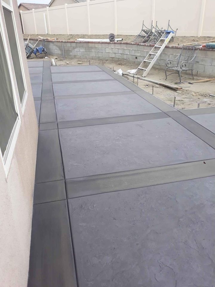 A freshly paved concrete patio next to a house, featuring smooth, gray concrete slabs with slightly raised edges. In the background, construction materials, a ladder, and an outdoor bench rest on a dirt area—work completed by a skilled concrete contractor in Albuquerque, NM.