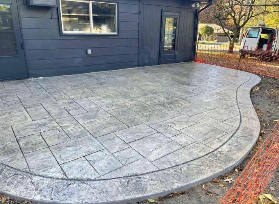 A newly installed gray concrete patio with a stamped, stone-like pattern graces the front of a dark-colored house. A window and door adorn the wall, while an open white van and orange safety netting linger in the background. For precision work like this, consider Quality Concrete NM for a free quote.