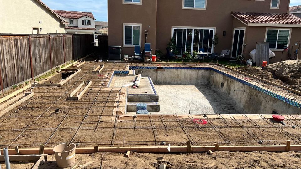 At an Albuquerque construction site, the backyard pool is taking shape with rebar and wooden framing in place. Skilled concrete contractors will soon create a sleek concrete pool deck. The area features mostly bare soil, while a two-story house stands prominently in the background.