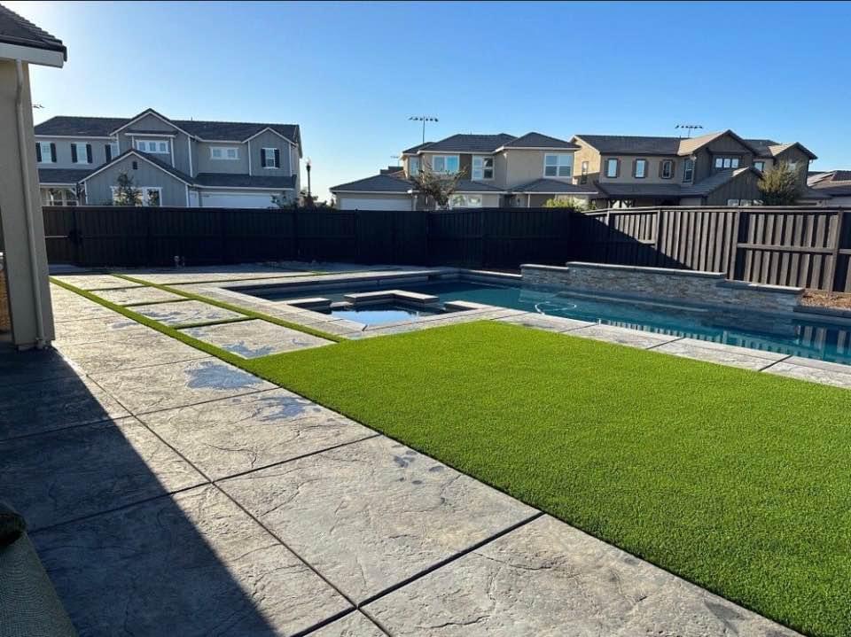 A modern backyard features a rectangular pool with an attached hot tub, expertly installed by Albuquerque's leading concrete contractor. The area is surrounded by artificial grass, stone pavement, and a dark wooden fence. In the background, several two-story houses lie under a clear blue sky.