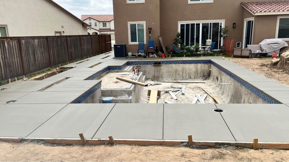 An unfinished backyard swimming pool surrounded by newly poured concrete needs. Construction materials and tools are visible inside the empty pool, highlighting Quality Concrete NM’s dedication to excellence. Albuquerque homes and patio furniture sit serenely in the background.